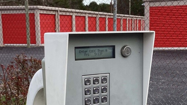 Photo of keypad to enter the storage facility. Behind it shows a chainlink fence and various drive-up storage units with orange doors